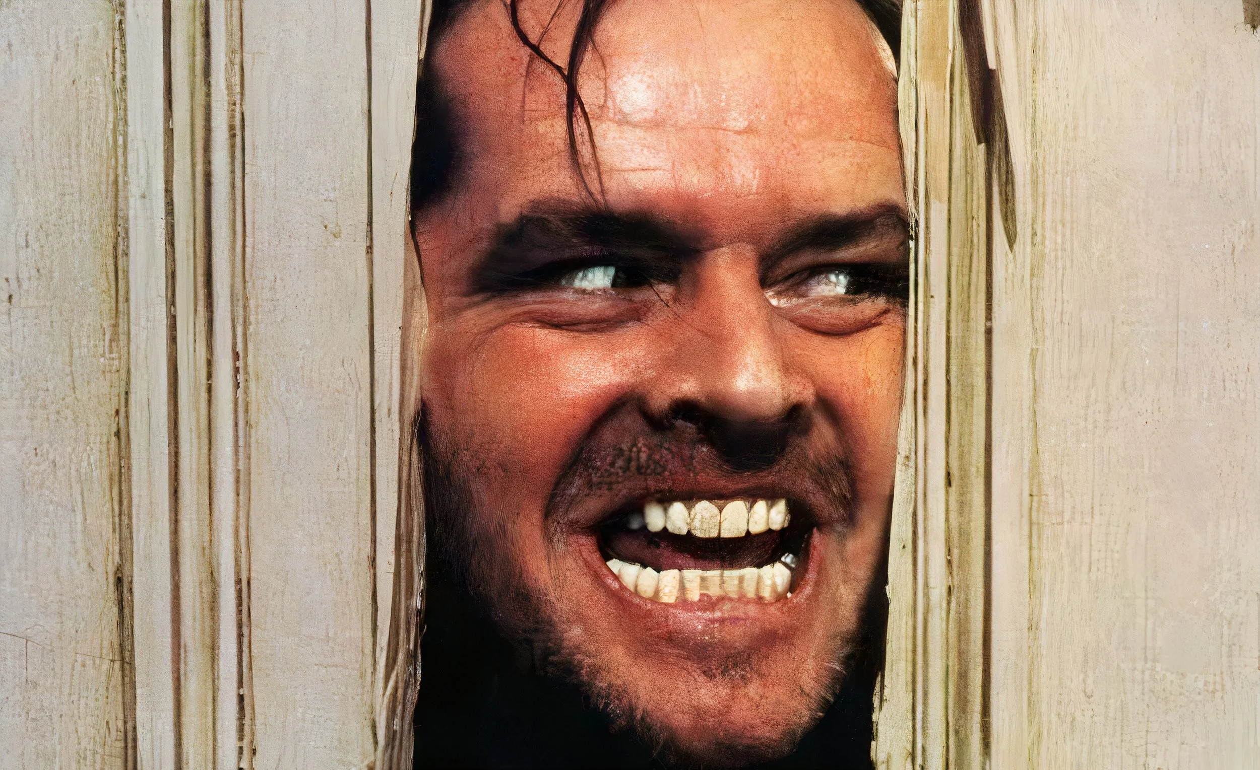 A man with dark hair and facial hair smiling mischievously through a wooden fence with gaps.