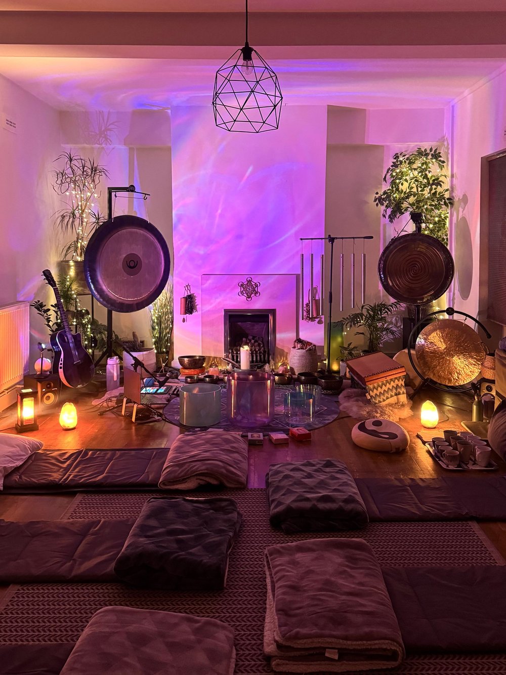 Meditation room setup with gongs, chimes, candles, cushions, and soft lighting for a tranquil ambiance. Plants and instruments are placed around a central area.
