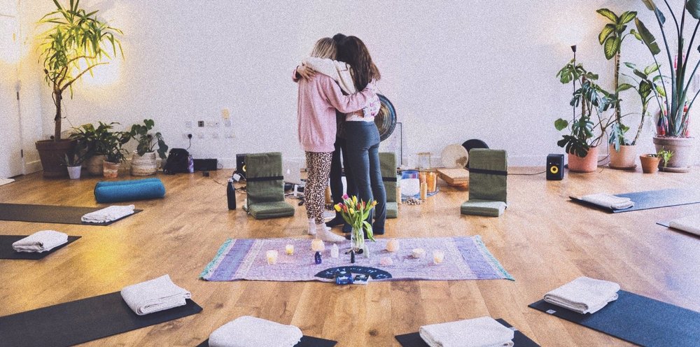 Three people hugging in a serene room with yoga mats, meditation cushions, plants, and candles.