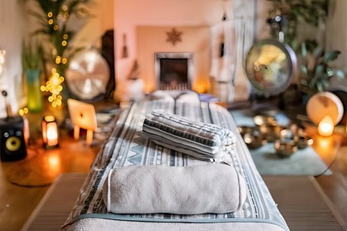 Calm, cozy home spa setting with candles, soft lighting, towels on a massage table, and blurred background items like a laptop, speaker, and decorative objects.
